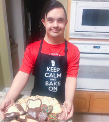 One of our newest Microgrant Recipients, Arturo Arias, smiling in the kitchen and holding a tray of his heart-shaped shortbread cookies dipped in chocolate. He's wearing an apron that says, "Keep calm and bake on". 