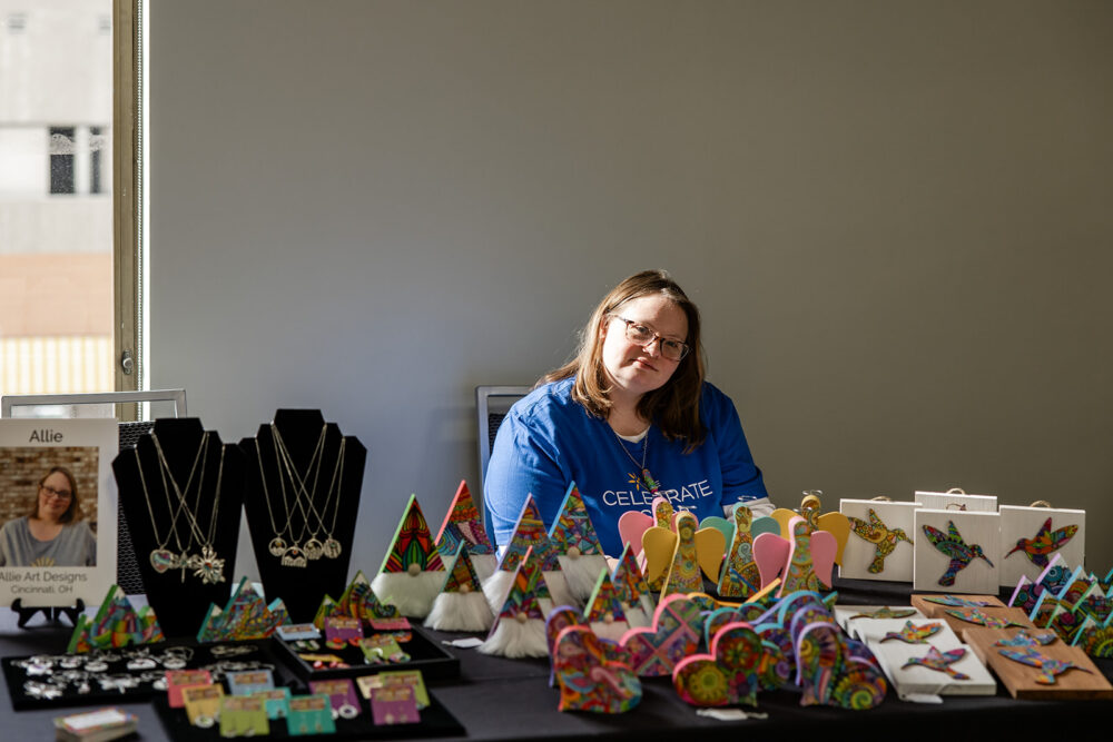 Entrepreneur Allie sitting at a table covered with her artwork at The Celebrate Shop
